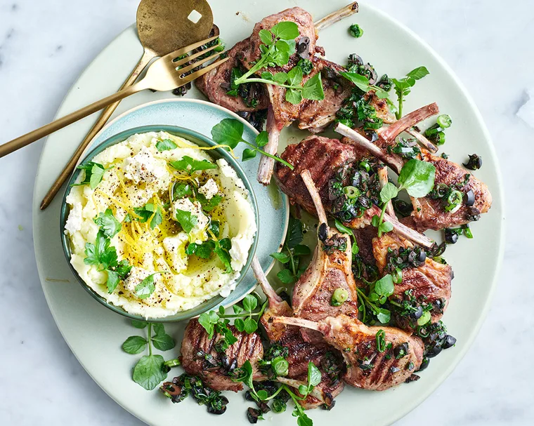 Lamb cutlets with feta skordalia and micro herbs on white plate with gold spoon and fork.
