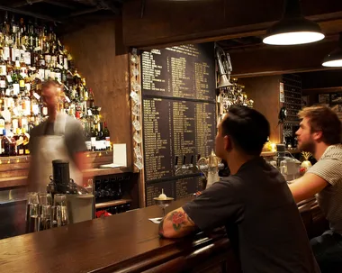 Photo of two men sitting at the bar and bartender blurred in motion in front of large whisky collection in back bar at Baxter Inn in Sydney, which is one of the best bars Australia that have had a lasting impact