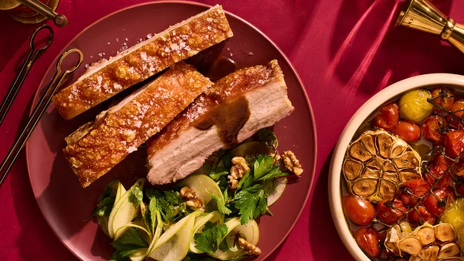 Twice-cooked pork belly roast with pickled apple salad and maple-glazed cabbage on red background.