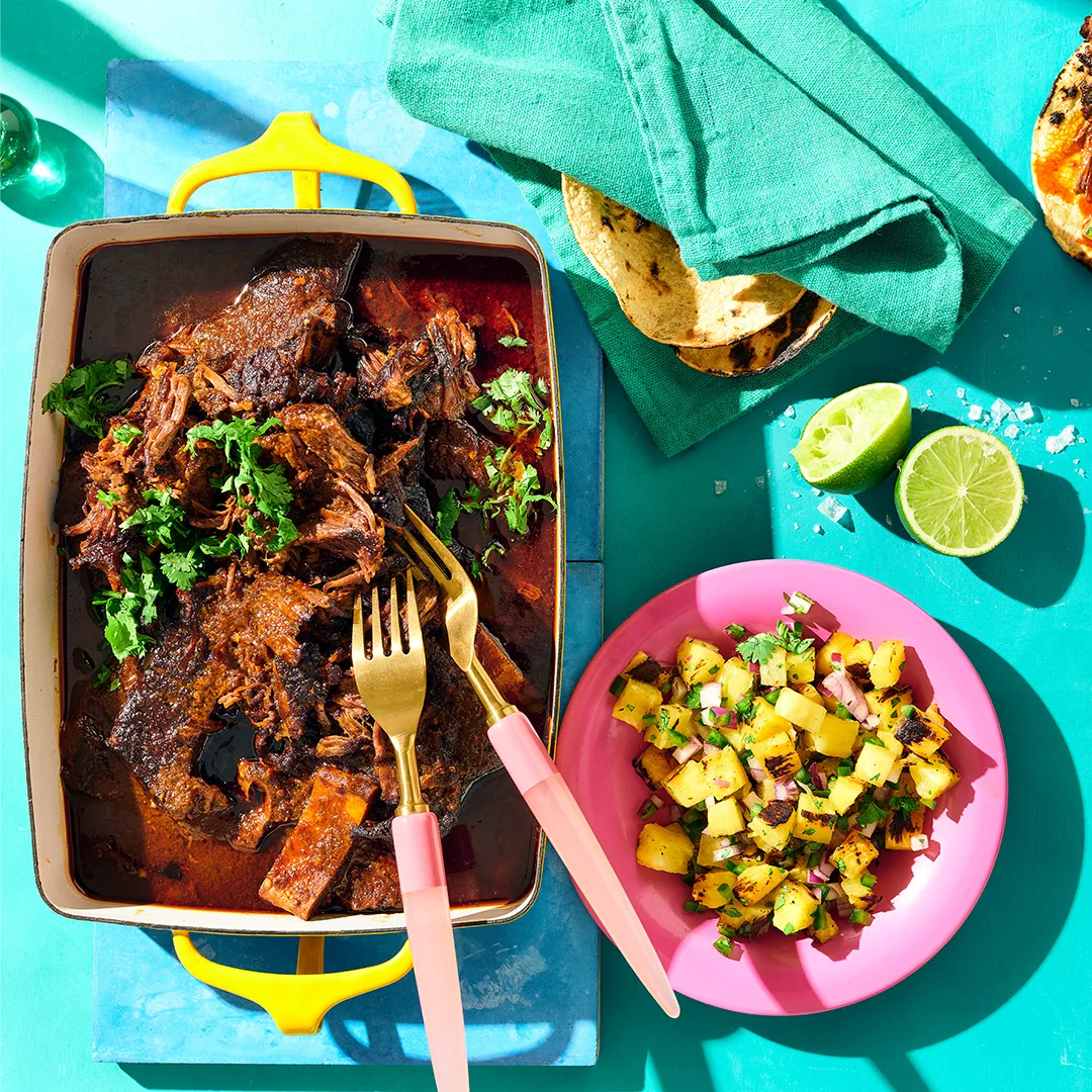 Birria tacos recipe with shredded beef short ribs in yellow baking dish in birria sauce, next to pink plate of charred pineapple salsa and corn tortillas on a birgh blue tablescape.