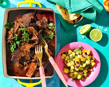 Birria tacos recipe with shredded beef short ribs in yellow baking dish in birria sauce, next to pink plate of charred pineapple salsa and corn tortillas on a birgh blue tablescape.
