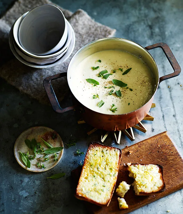 A pot of cauliflower soup sprinkled with sage, next to a chopping board holding one and a half slices of cheesy toast. Three stacked bowls sit to the top-left of the frame.