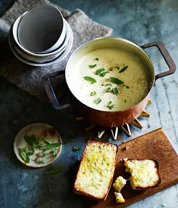 A pot of cauliflower soup sprinkled with sage, next to a chopping board holding one and a half slices of cheesy toast. Three stacked bowls sit to the top-left of the frame.