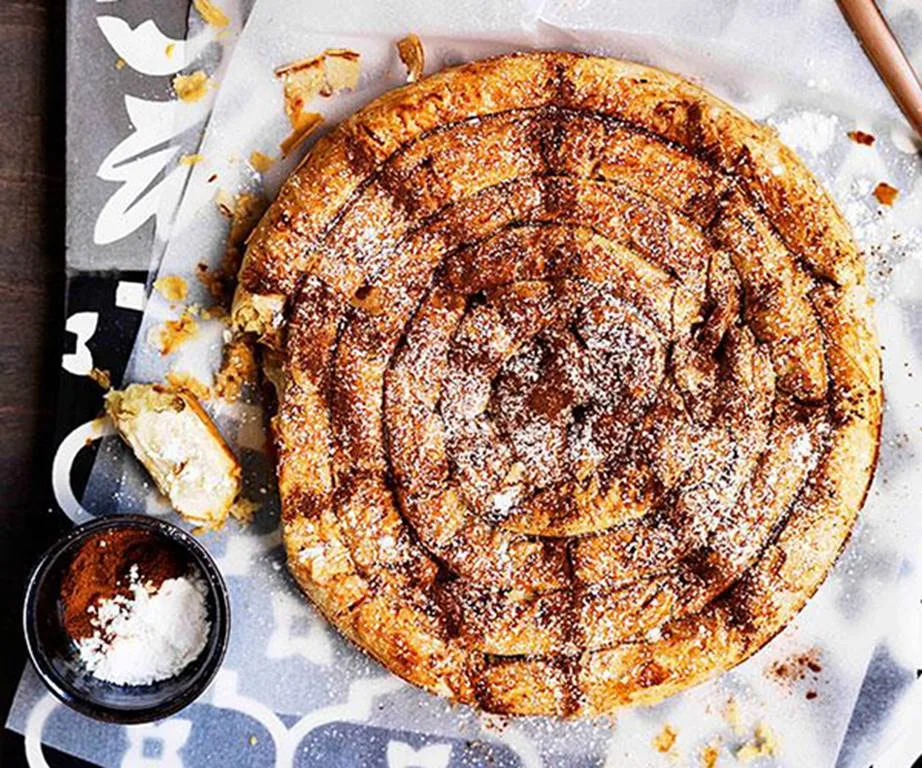 A spiral of baked pastry, dusted in icing sugar, on a piece of baking paper on a blue patterned placemat.