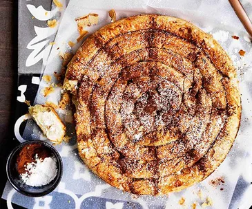 A spiral of baked pastry, dusted in icing sugar, on a piece of baking paper on a blue patterned placemat.