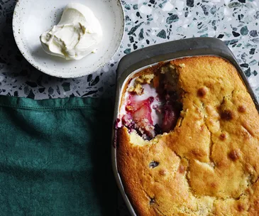 Apple and and berry pudding in a rectangle dish, with a scoop taken out of it, sitting on a green napkin, with a plate of vanilla ice-cream in the top-left