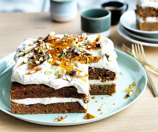 A square carrot cake, topped with cream-cheese icing and pistachios, on a eggshell-blue plate.