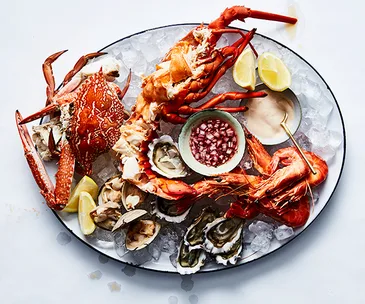 Over the top shot of a large white oval platter laden with seafood: a whole crab, a whole lobster, cooked prawns, clams, and shucked oysters. 