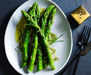 Five asparagus spears drizzled with butter, on a white oval plate, with a golden pepper shaker and black knife and fork on the side.