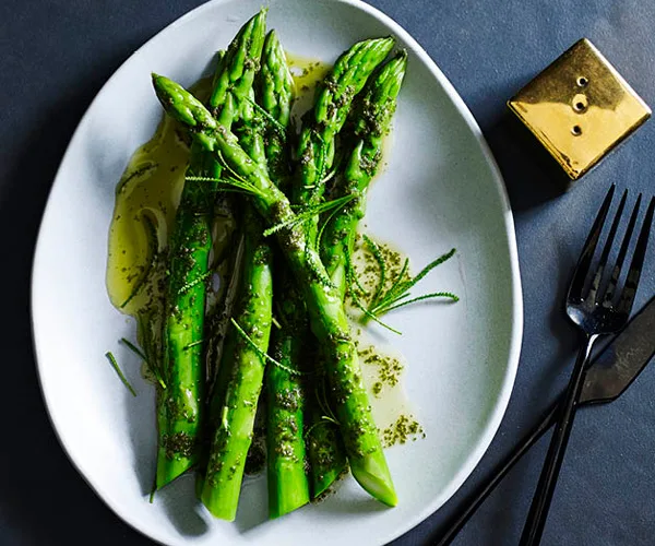 Five asparagus spears drizzled with butter, on a white oval plate, with a golden pepper shaker and black knife and fork on the side.