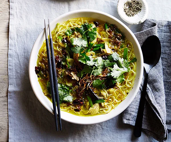 Overhead shot of a white bowl with turmeric soup, noodles and chicken, garnished with chopped coriander and fried shallots