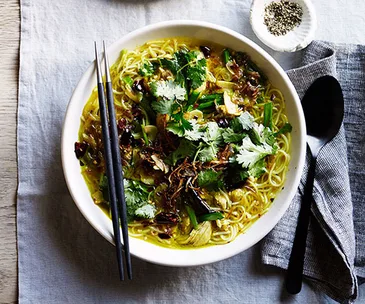 Overhead shot of a white bowl with turmeric soup, noodles and chicken, garnished with chopped coriander and fried shallots
