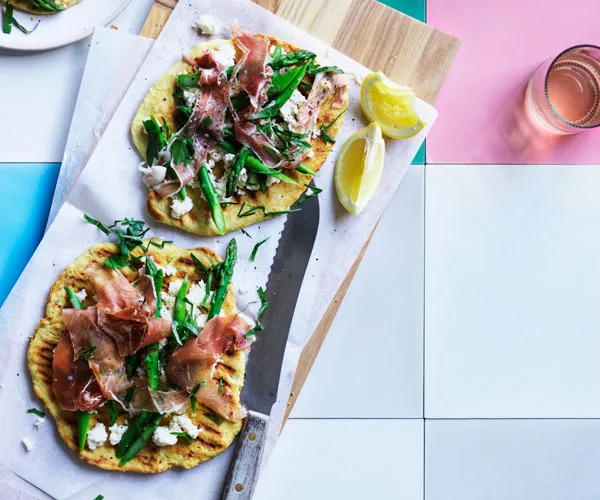 Two round grilled flatbread, topped with asparagus, prosciutto, and ricotta, on a rectangular wooden board wiht a bread knife and lemon segments, sitting on a pink, white and blue tiles.