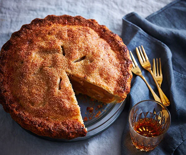 A round, crusty apple pie with a slice taken out of it, sitting on a light-blue linen tablecloth, with golden forks and a tumbler of amber-hued liquid to the side