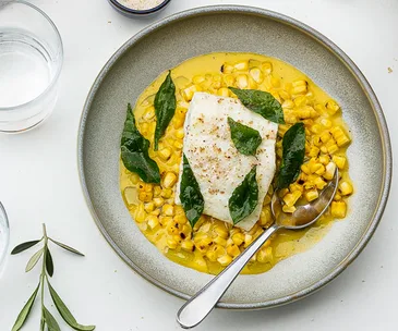 Bird's eye view image of grey ceramic bowl displaying yellow corn custard, white fish fillet and seven fried curry leaves, plus a silver spoon