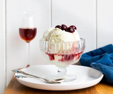 photo of wooden table and white wall with champagne flute filled with red liquid and a glass footed ice cream bowl with white ice cream, red liquid and topped with dark cherries in foreground