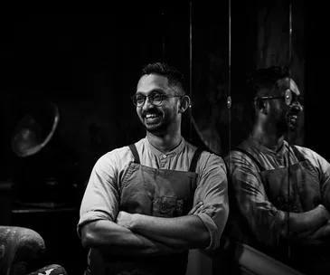 Black and white portrait of chef Rishi Naleendra leaning against a reflective wall.