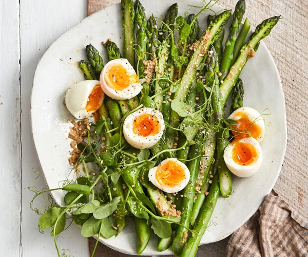 White plates with asparagus spears, halved soft-boiled eggs, and pea shoots, scattered with buckwheat.