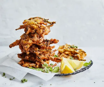Fried zucchini fritters with wedges of lemon, stacked on a plate lined with white baking paper, on a white background.