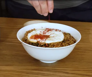 A white bowl holding jjapaguri noodles and a fried egg, with a hand sprinkling gochugaru Korean chilli flakes on top.