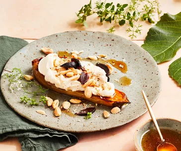 A thick slice of charred eggplant, topped with a thick white sauce and almonds, on a marbled grey plate, surrounded by a green linen napkin and green foliage.