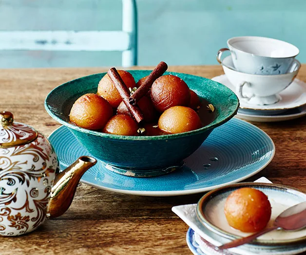A aqua-blue bowl holding spheres of gulab jamun in syrup, garnished with two cinnamon sticks