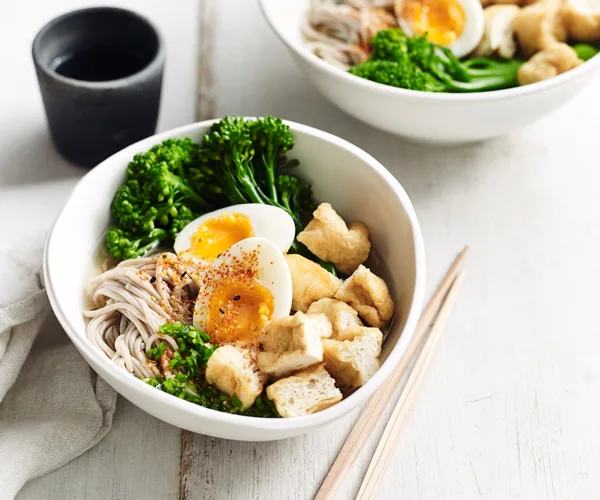 A white bowl with soba noodles, egg, tofy and broccolini, with a pair of wooden chopsticks on the side