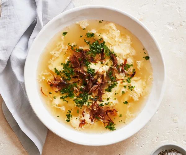 A white bowl holding a yellow-coloured egg-drop soup, scattered with fried shallots and chopped green herbs.