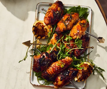 Skewers of barbecued chicken wings and lemon halves on a white rectangle tray, on an off-white marble table