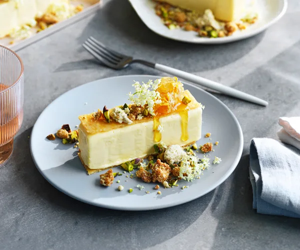 A rectangular block of light-green olive-oil ice-cream, covered with pistachio and honey, on a eggshell blue round plate, on a grey table.