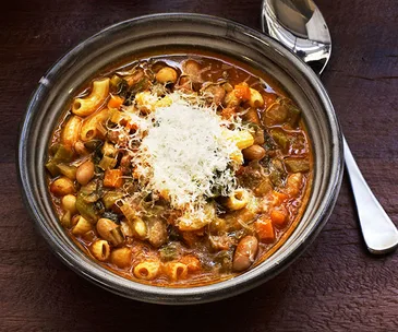 A minestrone soup with short pasta and vegetables, topped with grated parmesan, in a brown bowl. A silver spoon sits to the right. 