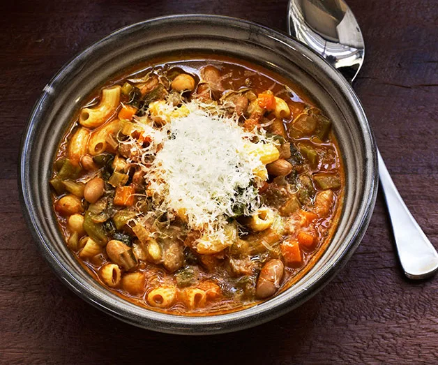 A minestrone soup with short pasta and vegetables, topped with grated parmesan, in a brown bowl. A silver spoon sits to the right. 