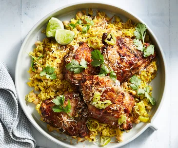 Close-up of a plate of roast chicken thighs on a bed of yellow pilaf, garnished with coriander leaves and lime wedges.