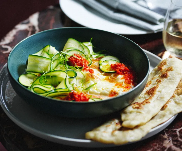 A dark green bowl with baked burrata, chilli sauce and slivers of zucchini, with some flatbread to the side.