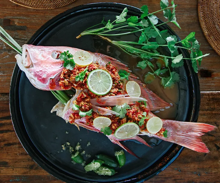 A whole steamed snapper on a black plate, garnished with sliced limes, crushed chillies and coriander sprigs.