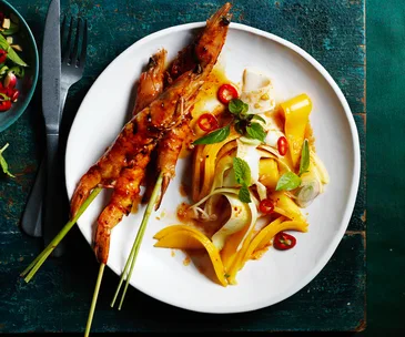 Three prawns on lemongrass skewers with strips of mango and coconut topped with chillies, mint, and shallots on a white plate.