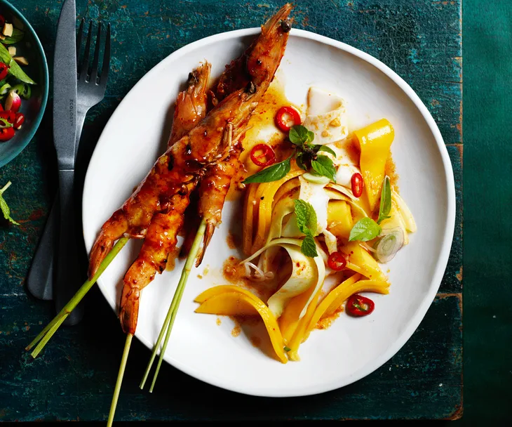 Three prawns on lemongrass skewers with strips of mango and coconut topped with chillies, mint, and shallots on a white plate.