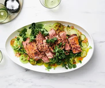Sliced grilled steak on a bed of sliced green tomato and honeydew melon salad. Topped with harissa butter and served on a white, egg shaped plate.