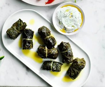 Dolmades wrapped in a silverbeet leaf served on a white platter and drixxled with olive oil. Tzatziki served on the side for dipping.