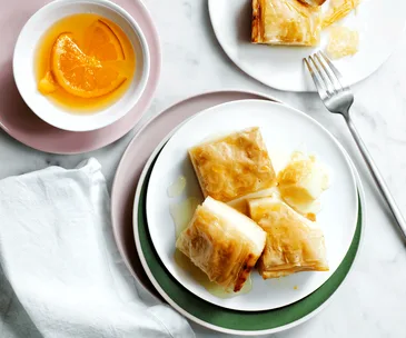 Three pieces of galaktoboureko stacked together on a white plate with orange syrup served in a dish on the side.