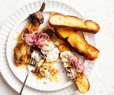 Whole smoky eggplant served on a white plate with three pieces of crusty bread and Greek yoghurt topped with red onion and sumac.