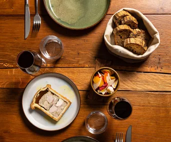 Bird's eye biew of wooden table displaying wine glasses and plates of terrine, pickles and bread
