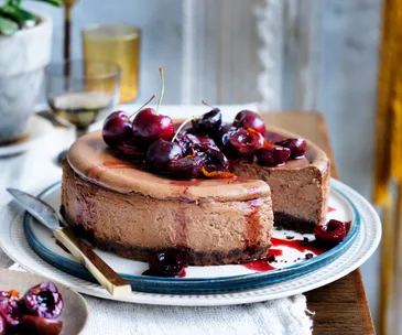 photo of table with white tablecloth and a blue-rimmed plate underneath a brown cheesecake topped with glossy cherries