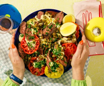 bird's eye biew image of two hands holding a blue plate topped with steak, sliced tomato, herbs and lemon