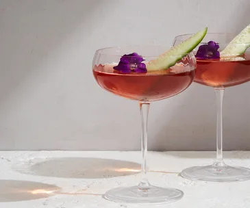 Two stemed glasses with Wallflower cocktail in them wth cucumber and purple flower garnish