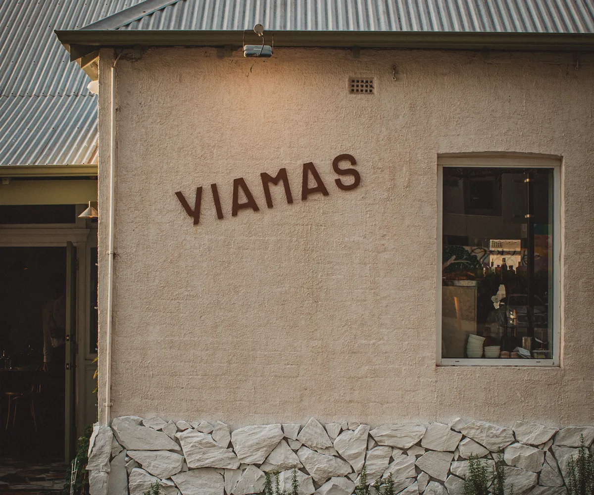 Exterior of stone-coloured building with sign reading 'YIAMAS' and a portrait window and metal roof