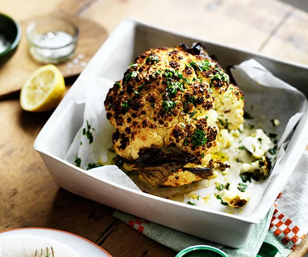 Whole head of roasted cauliflower, with a scorched top, scattered with parsley, in a square roasting tray.