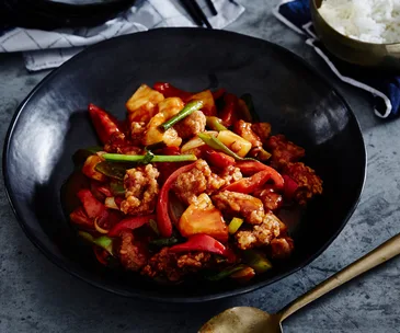 A black bowl holding sweet and sour pork with sliced capsicum and chunks of pineapple