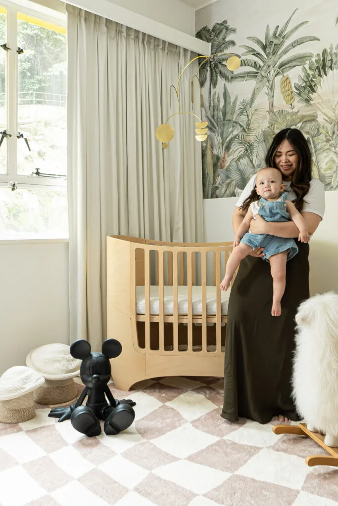 Woman holding baby in a neutral, jungle-themed nursery with a beige checkered floor rug.