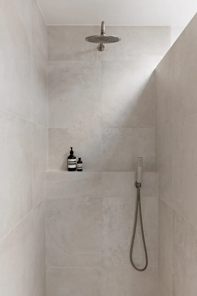 Shower in modern bathroom with large natural stone tiles and brass showerhead and skylight.